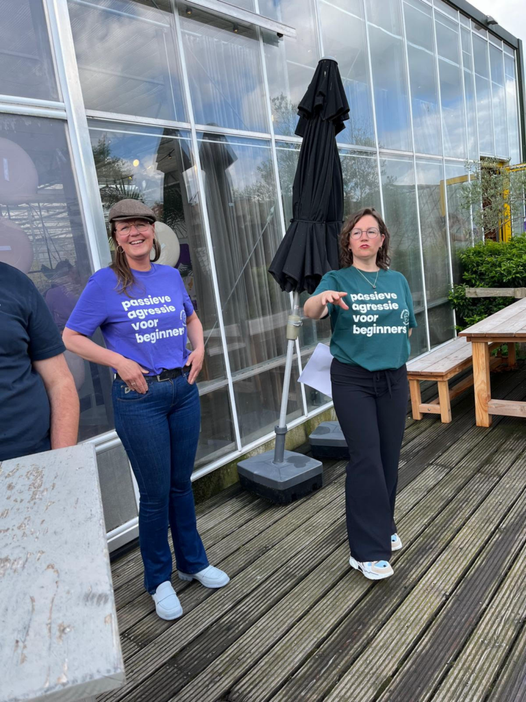 Je ziet Annelies en Sanne, ze staan buiten op een houten vlonder voor een kas. Annelies en Sanne hebben een t-shirt aan met daarop de tekst: passieve agressie voor beginners