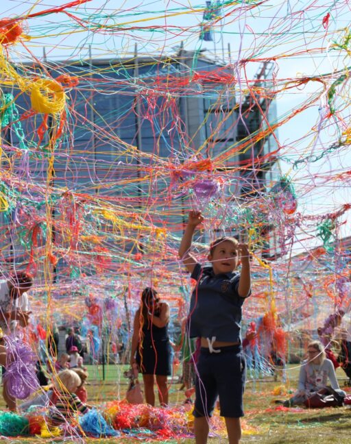 Je ziet mensen en kinderen onder een gigantisch web van gekleurde draadjes. Iedereen werkt mee aan het ophangen